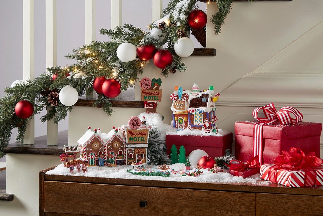 Gingerbread village display on a snowy tabletop with candy houses ornaments and red gift boxes under a pine garland on stairs