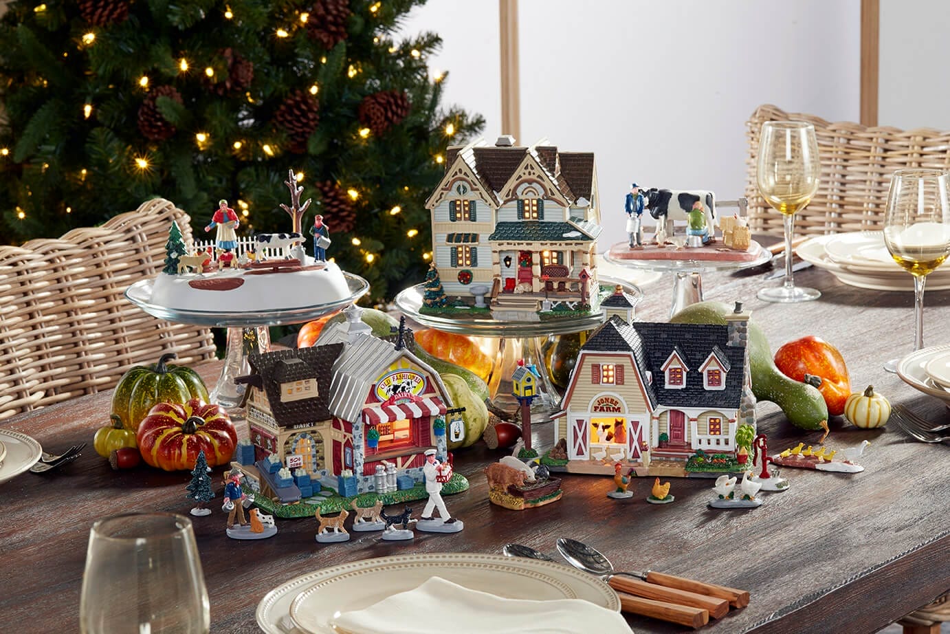 A festive tabletop display of miniature houses and figures arranged like a village, with pumpkins and a Christmas tree in the background on a dining table.