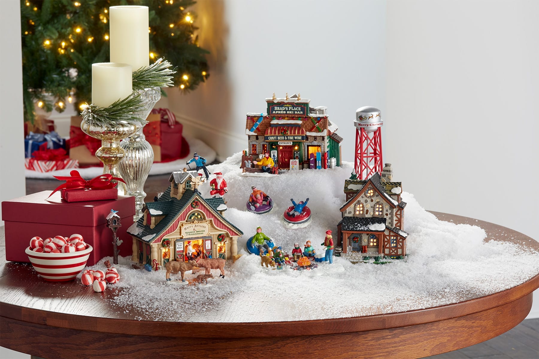 How to Pick a Village Theme Without Boxing Yourself In Lemax Collection Miniature Christmas village scene on a snow covered table with tiny houses figures gifts and a bowl of peppermint candies