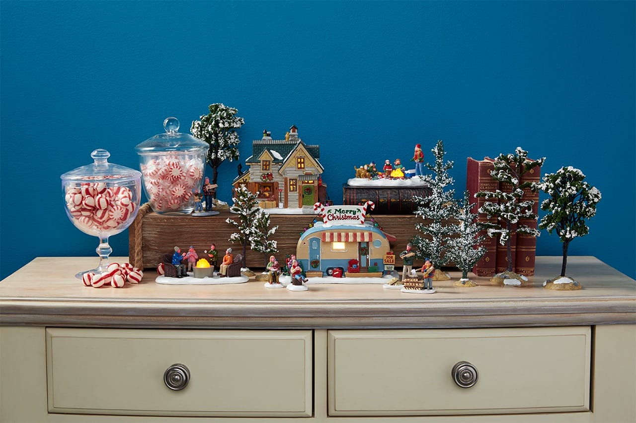 How to Pick a Village Theme Without Boxing Yourself In Lemax Collection Miniature Christmas village on a wooden dresser candy jars snow topped trees a lit house market stall and tiny shoppers